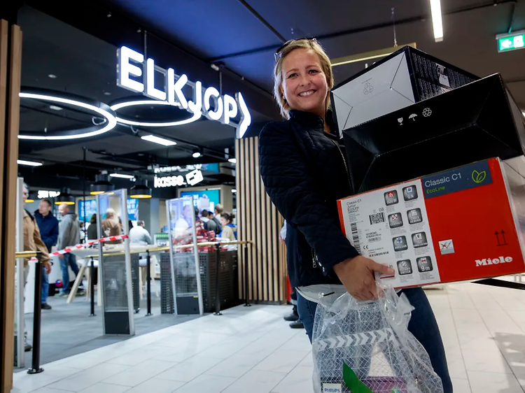 Kvinne kommer ut fra Elkjøp butikk og smiler med flere esker i hendende