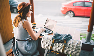 kvinne med hatt sitter på kafé med laptop og kaffe og smiler