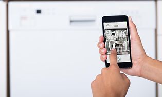 Hånd holder smarttelefon foran en oppvaskmaskin