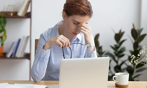 Kvinne sitter foran laptop med briller i hånda, indikerer hodepine eller slitne øyne