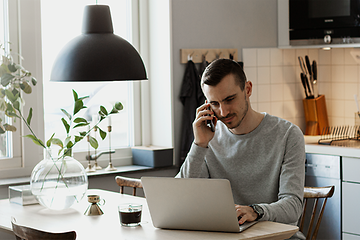 en mann jobber poå sin bærbare PC mens han snakker i smarttelefonen på et kjøkken