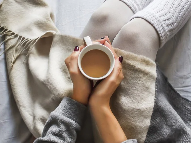 kvinne sitter i en seng med teppe over beina og kaffekopp i hendene
