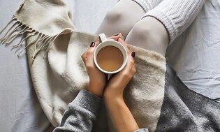 kvinne sitter i en seng med teppe over beina og kaffekopp i hendene