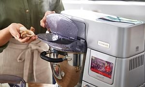 En kvinne er i ferd med å legge sopp i Kenwood Cooking Chef XL kjøkkenmaskinens bakeskål som har lokk