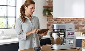 En kvinne som står med et nettbrett og bruker Kenwood World-appen ved siden av en Kenwood Cooking Chef XL kjøkkenmaskin