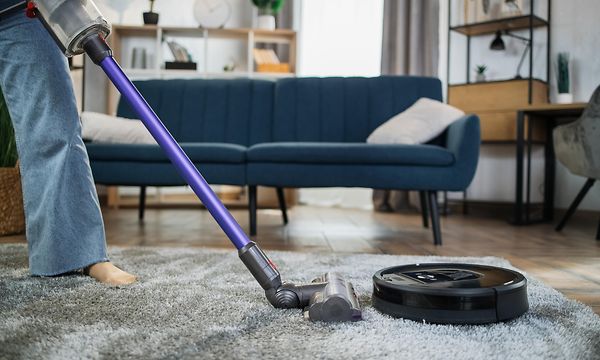 En person som bruker en trådløs støvsuger i en stue ved siden av en svart robotstøvsuger