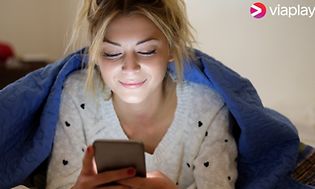 Smilende ung kvinne under teppe som ser på smarttelefon med Viaplay logo