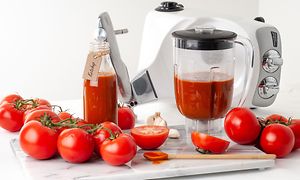 En Assistent blenderbeholder med tomatjuice med tomater omkring og en Ankarsum kjøkkenmaskin bak