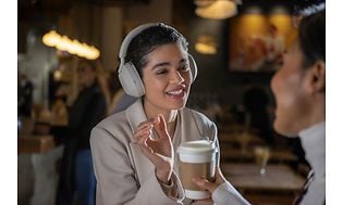 En kvinne har på seg hvite Sony WH1000XM5-hodetelefoner på en kafé og tar imot en kaffe