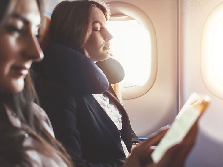 To kvinner slapper av på et fly, en med nakkepute og den andre ser på telefonen sin