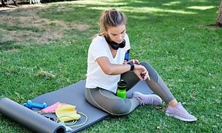en kvinne sjekker smartklokken sin mens hun sitter utendørs på en yogamatte
