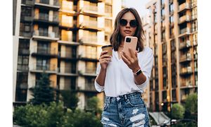 Kvinne med solbriller holder en kaffekopp og bruker en smarttelefon