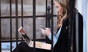 Ung kvinne som sitter ute og ser på mobilen med macbook på fanget