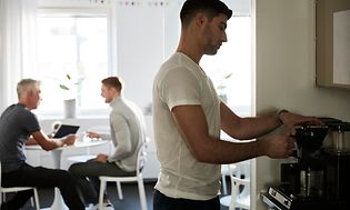 mann lager kaffe på en Moccamaster med to menn sittende i bakgrunnen