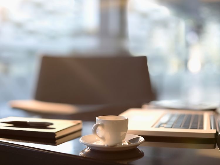 Kaffekopp med tefat på en pult foran macbook og skrivesaker