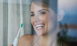Smilende kvinne ved vindu som holder en elektrisk tannbørste
