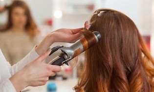 Hairdresser makes curls in the hair of a woman with long red hair in a beauty salon.