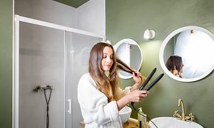 Woman straightening her long hair with hair straightener in bathroom