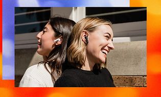 To jenter har hvert deres par JLB True Wireless ørepropper i ørene og står rygg mot rygg og smiler