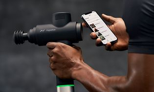 En mann som holder en Hyperice Hypervolt massasjepistol og telefon med Hyperice-appen