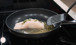 A person frying a fish on a pan with a plastic spatula