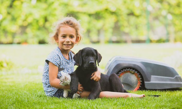 oung girll holding a puppy ona a lawn with a robot lawn mover beside them