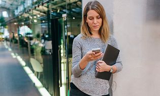 Kvinne som står i kontorkorridoren og sjekker telefonen mens hun holder en bærbar datamaskin i den andre armen
