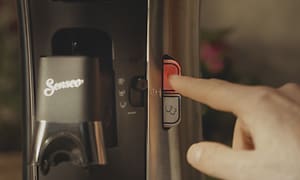 Nærbilde av en hånd som trykker på på-knappen på en Senseo Select-kaffemaskin