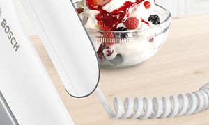 Nærbilde av Bosch StyLine-håndmikser sin strømkabel og en dessert i bakgrunnen
