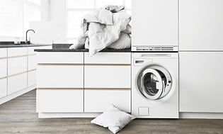 Hvitt vaskerom med frontmatet vaskemaskin og en haug med tøy på toppen av benken