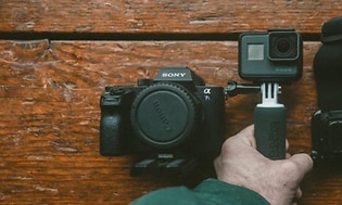 SLR-kamera og actionkamera i hånd med tilbehør på trebord
