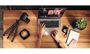 person redigerer vlogg på mac med kaffe, kamera, stativ og skrivesaker på bordet