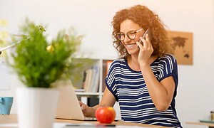 kvinne i kontorlandskap snakker i smarttelefon foran laptop