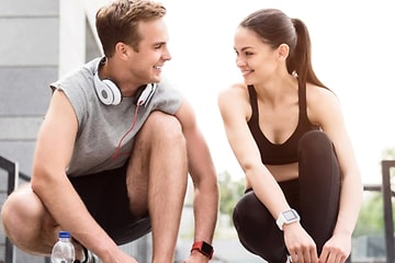 mann og kvinne i sportsklær, med headset og smartklokke, ser på hverandre mens de knytter sko