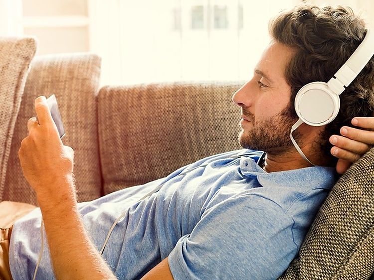 Mann ligger på sofa med smarttelefon i hånden og headset på hodet