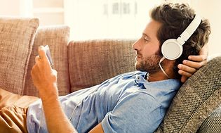 Mann ligger på sofa med smarttelefon i hånden og headset på hodet