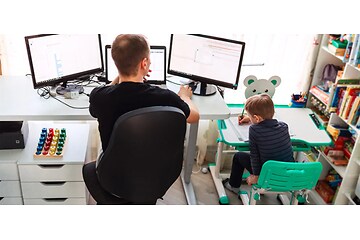 Mann sitter foran tre skjermer på hjemmekontor på barnerom med liten gutt ved siden av seg