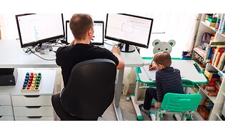 Mann sitter foran tre skjermer på hjemmekontor på barnerom med liten gutt ved siden av seg